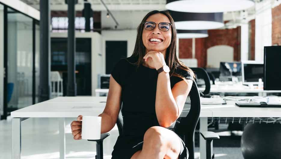 Happy young businesswoman feeling cheerful in a modern office. Young businesswoman smiling while sitting alone on a chair. Creative businesswoman enjoying her coffee break at work.